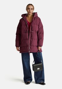Donna con giacca a vento viola con cappuccio, jeans a gamba larga, scarpe nere e una borsa nera, davanti a uno sfondo neutro.
