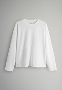 Ein schlichte, weiße Langarmshirt mit Rundhalsausschnitt hängt vor einem hellgrauen Wandhintergrund.