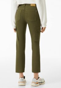 Osoba v olivově zelených cargo kalhotách s zadními a bočními kapsami, v kombinaci s bílými teniskami a bílým dlouhým rukávem, pohled zezadu.