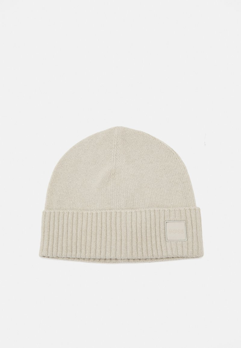 Bonnet tricoté beige clair avec revers côtelé plié et petite écusson carré sur le revers, posé sur un fond blanc uni.