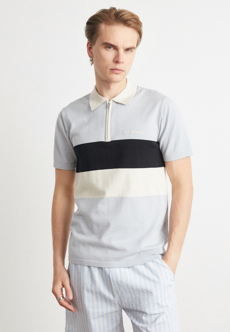 Polo de manga curta com fecho de zíper em cinza claro, com riscas horizontais em azul-marinho e crème. Apresenta um tecido suave e um bordado subtil no peito.