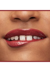 Rossetto rosso intenso su labbra piene con un finish lucido, che mette in evidenza denti bianchi e uniformi. Texture della pelle morbida attorno all'area della bocca.