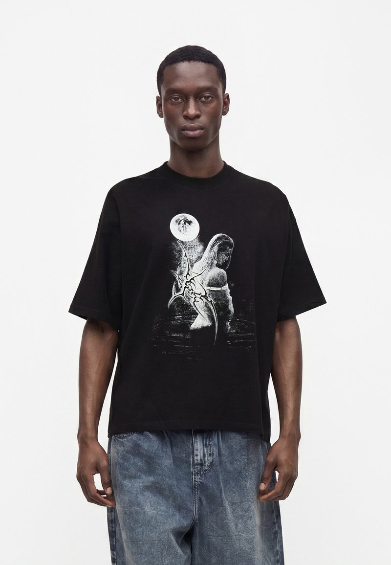 Άνδρας που φοράει μαύρο υπερμεγέθες t-shirt με λευκό γραφικό μιας γυναίκας και πανσέληνο, συνοδευόμενο από χαλαρά, ξεβαμμένα μπλε τζιν, στέκεται μπροστά σε απλό φόντο.