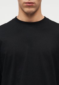 Μαύρο βαμβακερό μπλουζάκι με στρογγυλή λαιμόκοψη, κοντά μανίκια και λεία, απλή υφή, χωρίς σχέδια ή ορατά εξαρτήματα.