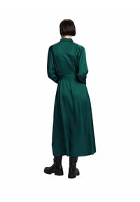 Longue robe en satin vert avec un col, une ceinture à la taille et un design fluide. Portée avec des bottes noires. Texture lisse et détails minimaux.