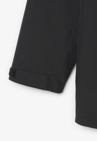 Giacca impermeabile nera con una texture liscia, chiusura a velcro sui polsini e marchio in rilievo sulla manica. Vestibilità su misura, design minimalista.