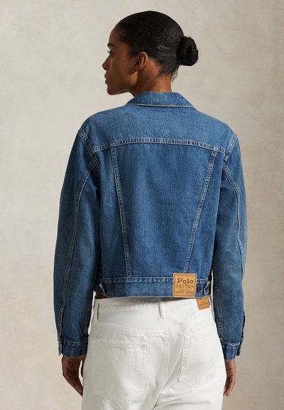 Donna con i capelli raccolti in uno chignon, che indossa una giacca di jeans blu e pantaloni bianchi, vista di spalle su uno sfondo neutro uniforme.