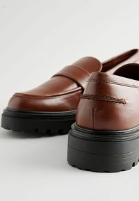 Mocasines de cuero marrón con una suela negra estriada, que presentan un upper suave y detalles de costura en el empeine y la parte trasera.