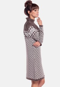 Kobieta w brązowo-białej dzianinowej sukience w wzory z golfem, stojąca bokiem z ręką przy brodzie, na jednolitym tle.