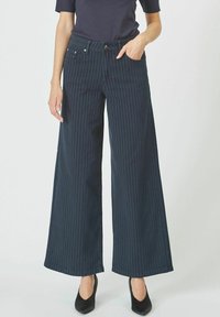 Marineblaue weit geschnittene Hose mit weißen vertikalen Streifen, hoher Taille, vorderen Taschen und einem dezenten Glanz. Kombiniert mit schwarzen Absätzen.