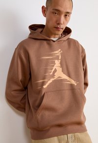 Sweat à capuche marron avec une finition texturée. Il présente un grand logo beige clair à l'avant et une poche kangourou. Coupe décontractée.