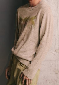 Pull beige à texture particulière, avec un motif fané de palmier en vert et marron. Manches longues avec poignets côtelés, porté avec un pantalon de couleur claire.