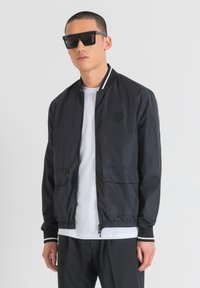 Giubbotto bomber nero con una texture liscia, caratterizzato da accenti a strisce bianche sul colletto e sui polsini, e due tasche frontali. Indossato sopra una camicia bianca.