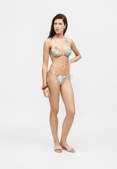 Femme debout en bikini string coloré à motifs avec bracelets et sandales beige à enfiler sur fond blanc uni.