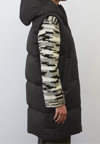 Svart ärmlös puffer-väst med huva, med en slät textur och en chunky stickad tröja i vitt och svart mönster under.