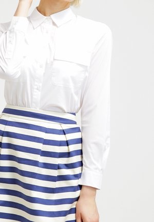 Kvinde iført en hvid skjorte med knapper og brystlomme samt et højtaljet skørt med horisontale blå og hvide striber.