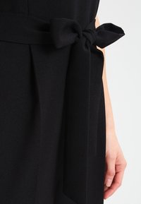 Svart tygklänning med ett knytbälte vid midjan, bredvid en avslappnad hand på höger sida.