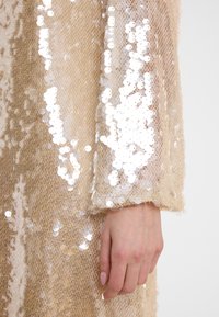 Vêtement pailleté avec une base beige clair, orné de sequins ronds brillants et réfléchissants. La manche est ample, révélant une main avec des ongles manucurés.