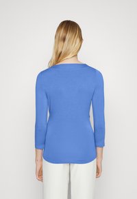 Mujer con cabello largo y rubio, vistiendo una blusa de manga larga azul y pantalones blancos, de pie de espaldas frente a un fondo plain.