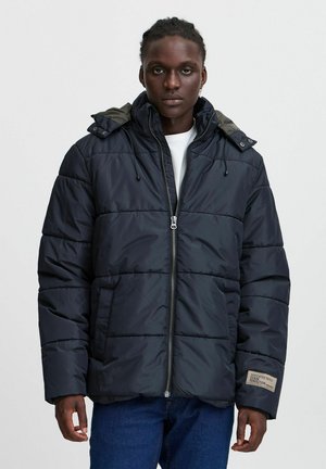 Puffer jacket in dark navy with gray hood, zip closure, and front pockets. Features horizontal quilting and a side label with text.