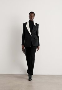 Black tailored blazer with a white peak lapel, paired with black fitted pants and a black turtleneck; model holds a sleek black clutch.