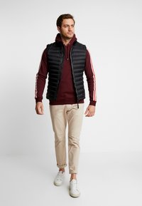 Black puffer vest over a maroon hoodie with white drawstrings, paired with beige pants and white sneakers. Simple design, relaxed fit.