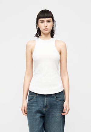 Mujer joven de cabello oscuro que lleva una blusa blanca sin mangas y jeans azules sueltos, de pie contra un fondo claro y liso.