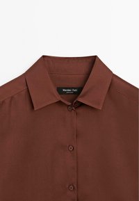 Chemise marron boutonnée avec col pointu, trois boutons visibles et une étiquette noire indiquant Massimo Dutti à l'intérieur de l'encolure.