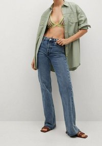 Chemise verte surdimensionnée, bralette rayée cropped, jeans bleus évasés taille haute et sandales marron. Tissu léger, coupe décontractée.