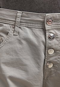 Pantalones de color gris claro con una textura suave, que cuentan con una bragueta de botones, cinco botones y bolsillos frontales. Detalles de costura a lo largo de los bordes.