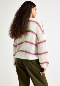 Krémový pletený svetr s růžovými a zelenými pruhy, s volným střihem a mírně nadrozměrnými rukávy, v kombinaci s olivově zelenými kalhotami.