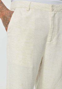 Pantalons en lin beige clair présentant une finition texturée discrète, des poches avant et une fermeture à bouton à la taille.