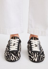 Zapatos slip-on con estampado de cebra en blanco y negro, con cordones blancos y un parche de cuero negro que presenta un logotipo dorado, vistos desde arriba.