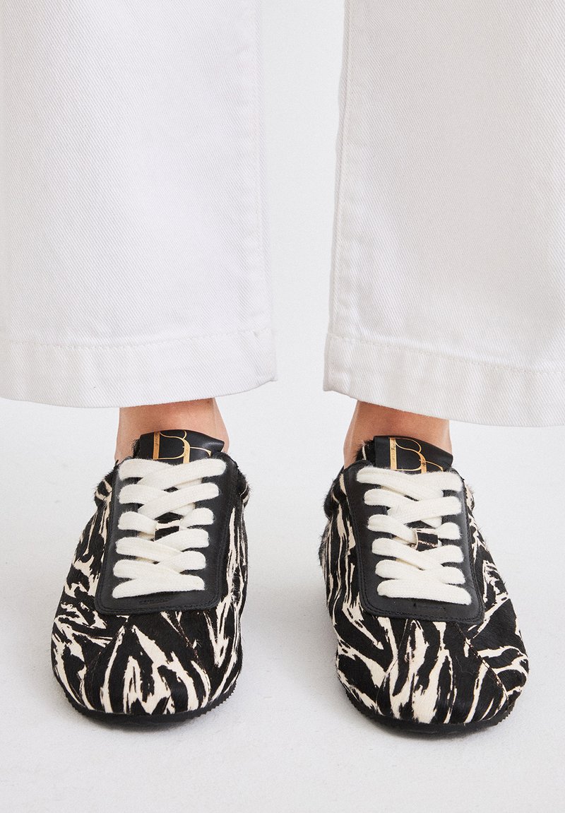 Zapatos slip-on con estampado de cebra en blanco y negro, con cordones blancos y un parche de cuero negro que presenta un logotipo dorado, vistos desde arriba.