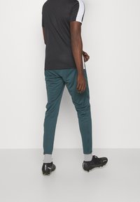 Zwarte sportshirt met witte accenten, gecombineerd met teal atletiekbroek en zwarte voetbalschoenen. De broek heeft een slank, taps toelopend ontwerp.