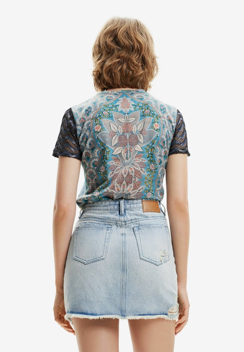 Kvinna med kort vågigt hår som bär en blommig blå topp med spetsärmar och en ljus denimkjol med fransar sedd bakifrån.