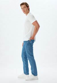 T-shirt bianca a maniche corte, jeans in denim blu e sneaker bianche. Il modello è in profilo laterale su uno sfondo neutro.