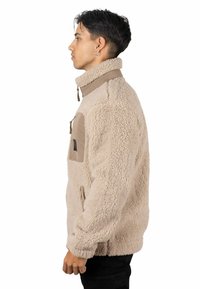 Chaqueta de forro polar beige con cuello alto, bolsillo en el pecho con cremallera y paneles de tela lisa en contraste. Material suave y texturizado con un ajuste relajado.