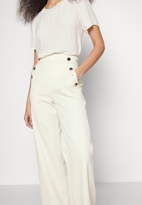 Pantalons larges de couleur crème avec une taille haute, présentant des accents de boutons sur les poches latérales et un tissu lisse et ajusté.