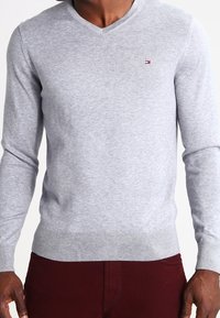 Grau gestrickter Pullover mit V-Ausschnitt und langen Ärmeln. Mit einem kleinen Logo auf der Brust sowie gerippten Bündchen und Saum.
