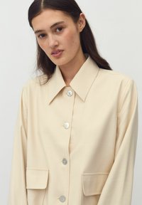 Beige takki, joka on valmistettu sileästä kankaasta, jossa on suuri kaulus, kaksi etutaskua ja hopeiset napit keltaisilla langoilla.