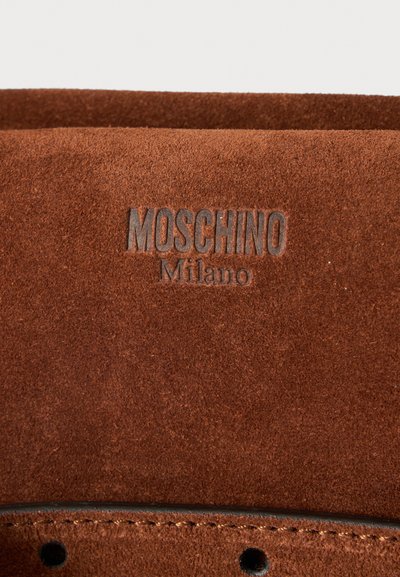 Surface en daim marron avec texte embossé ""MOSCHINO Milano"" au-dessus d'un bord en cuir cousu avec deux trous.