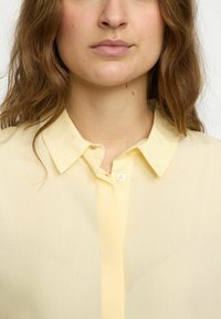 Light yellow button-up shirt with a soft texture, pointed collar, and a single button placket at the neck, displayed against a neutral background.