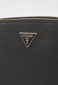 Mustmust tekstuuriga must rahakott, millel on kuldne tõmblukk, ning sellel on kolmnurkne must logoplekk, millel on kuldsete tähtedega "GUESS USA" ja küsimärk.