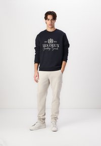 Sudadera de cuello redondo de color navy con el logo blanco impreso "LES DEUX Sporting Goods" combinada con pantalones cargo de color beige claro y zapatillas blancas.
