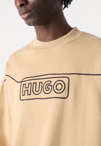 Beige pulóver kerek nyakkivágással, sötét hímzett "HUGO" logóval és sima, textúrált anyagbéléssel.