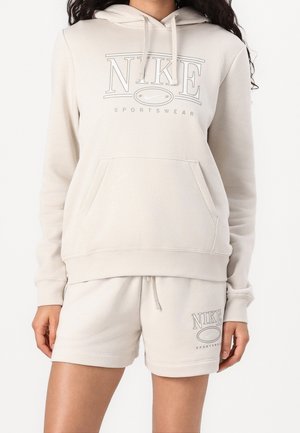 Femme portant un hoodie Nike beige clair et un short assorti avec les logos Nike Sportswear sur les deux pièces.