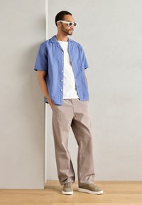 Camicia a maniche corte azzurra con bottoni sopra una t-shirt bianca, abbinata a pantaloni beige e sneakers grigie, su uno sfondo neutro.