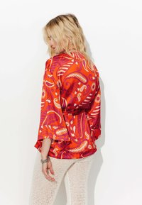Blouse à motifs rouges et orangés avec des manches flottantes et une ceinture à la taille, associée à un pantalon blanc texturé et léger.
