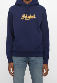 Person som bär en marinblå huvtröja med gula "Ralph"-bokstäver och ljusblå denimjeans, stående mot en neutral bakgrund.